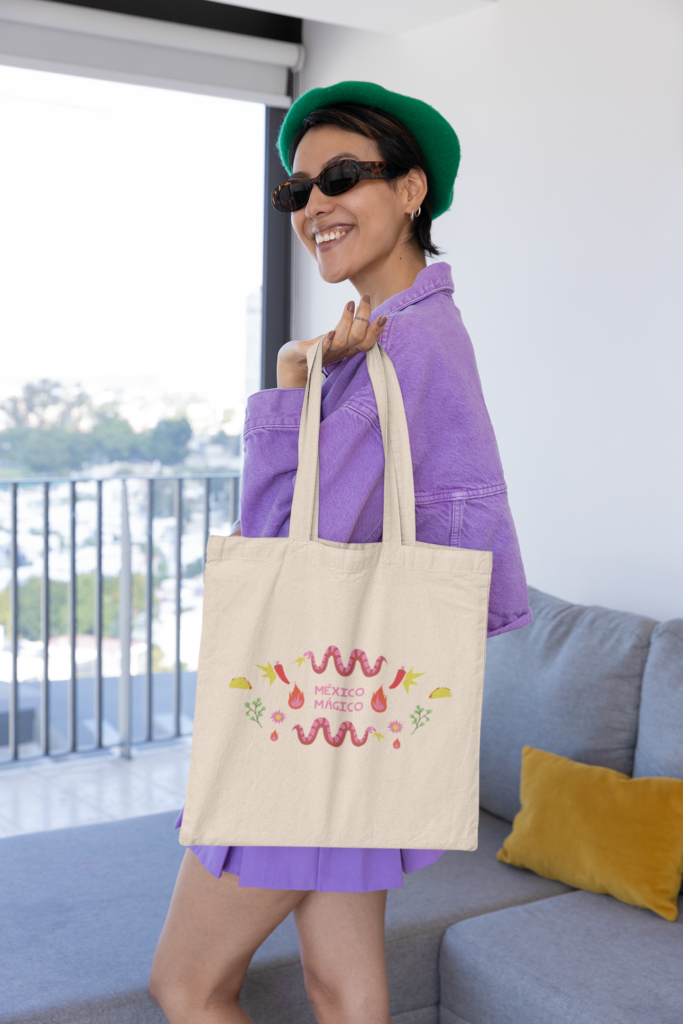 Una mujer que en la mano izquierda lleva una tote bag con el nombre de méxico mágico en colores mexicanos como rosa, rojo, amarillo. Esta sonriendo.